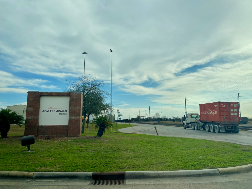 APM Terminals Mobile entrance sign, with a loaded container truck driving by and a crane in the background