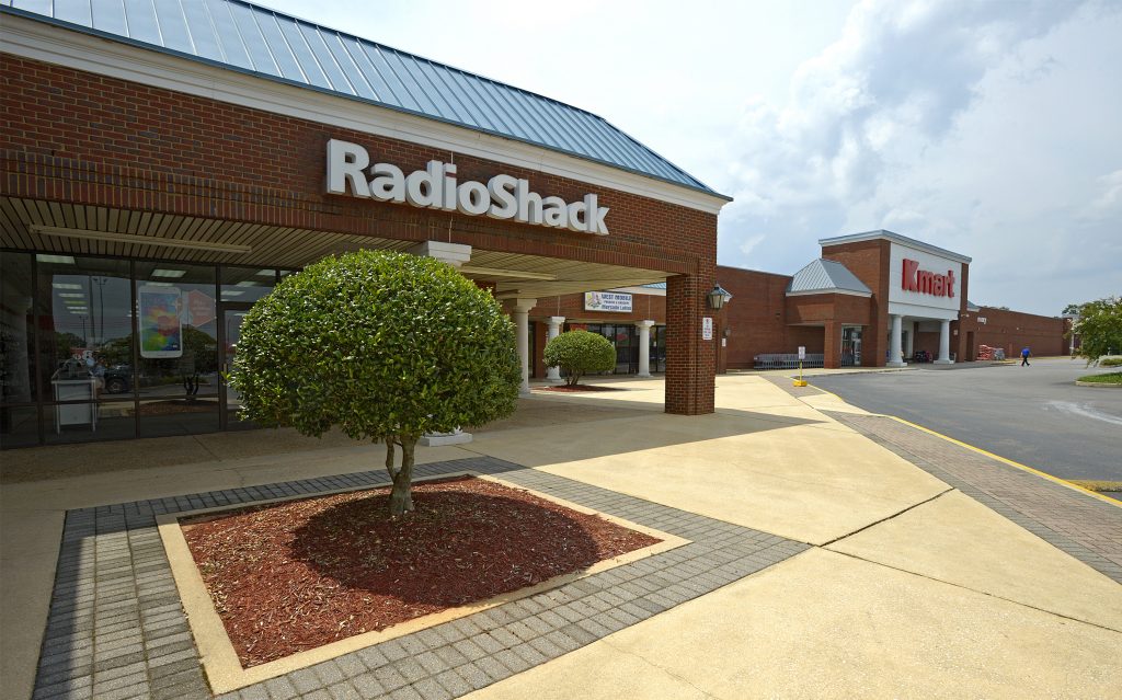 Retail center featuring a nicely landscaped bush and well-maintained sidewalk, with RadioShack and KMart in the background. 